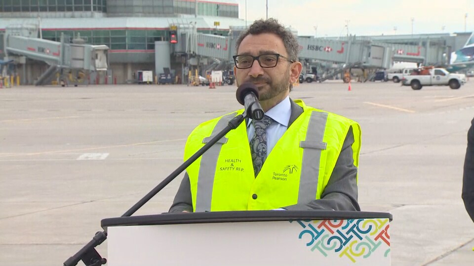 Omar Alghabra, vêtu d'un gilet de sécurité, répond aux journalistes sur le tarmac de l'aéroport Pearson.