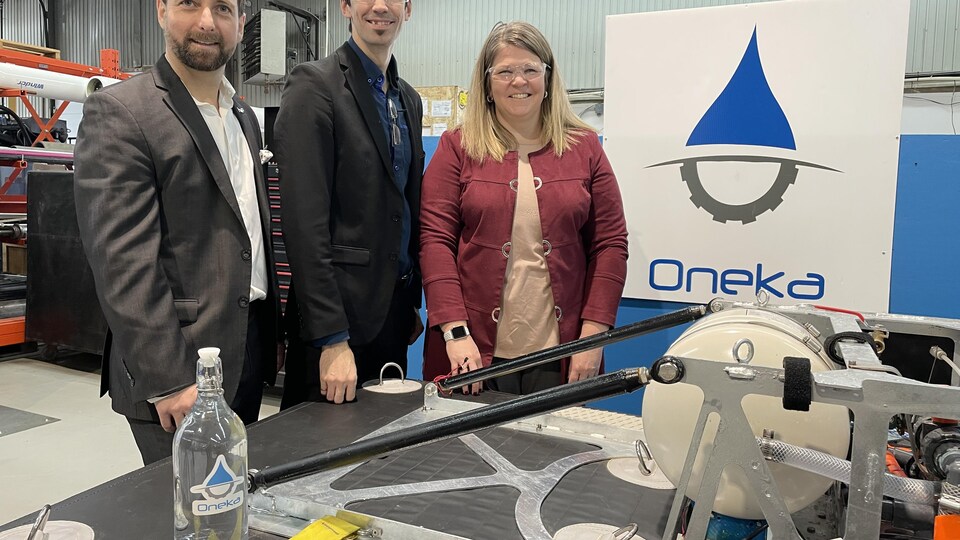 L'équipe d'Oneka Technologies, deux hommes et une femme, sourient devant une affiche de leur entreprise. 
