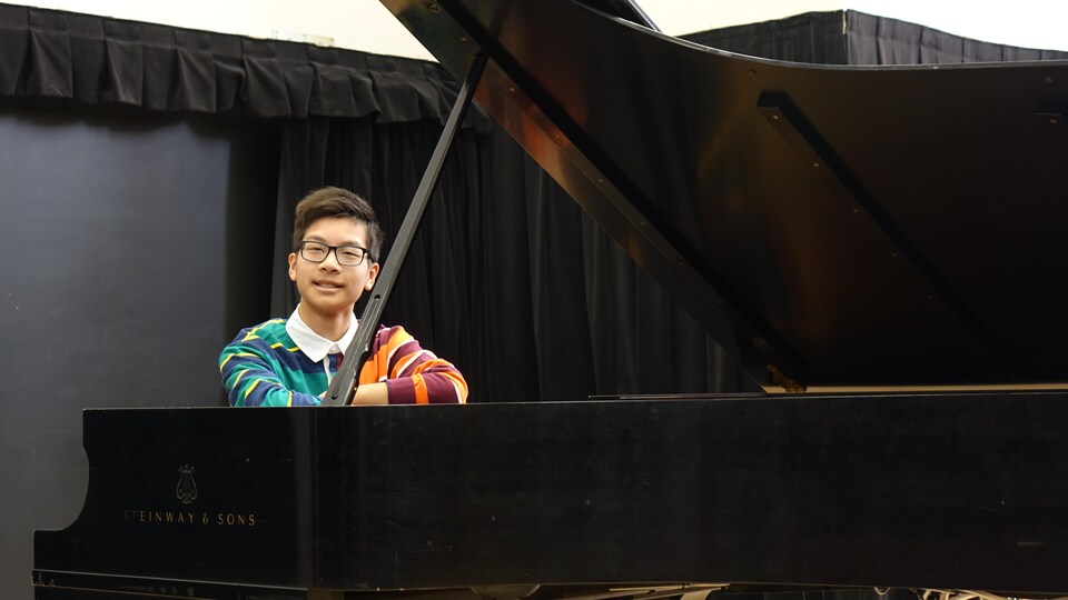 Un pianiste de 15 ans d'origine chinoise est assis au piano. Il porte un chandail rayé aux couleurs vives. Il a des lunettes.