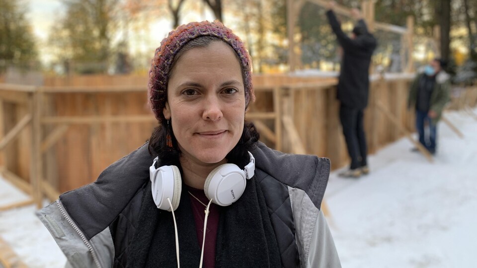 Une femme souriante avec un casque d'écoute autour du cou devant une patinoire extérieure.