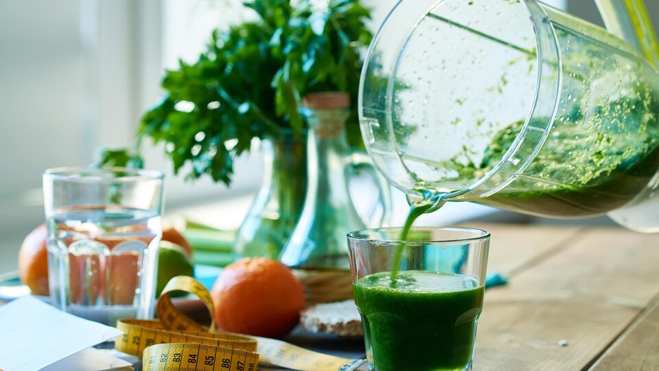 Un pichet qui verse un jus vert dans un verre sur une table entouré d'autres aliments et d'un ruban à mesurer.