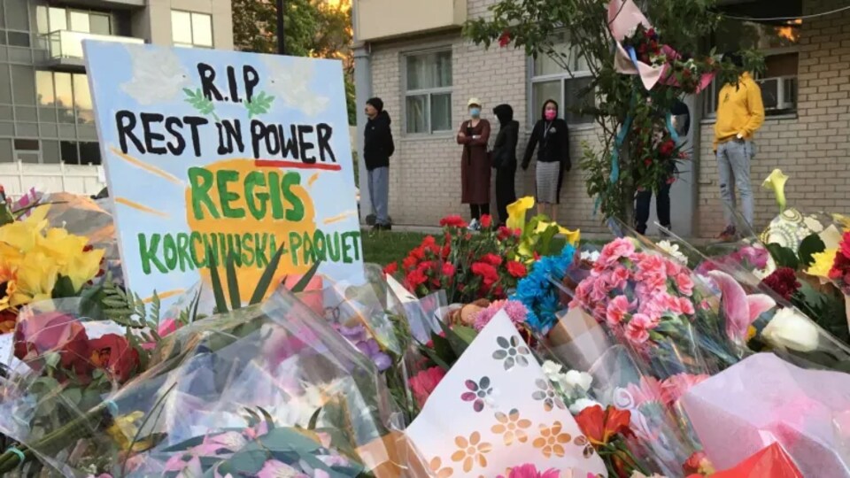 Une pancarte en mémoire de Regis Korchinski-Paquet est entourée de bouquets de fleurs devant un immeuble.