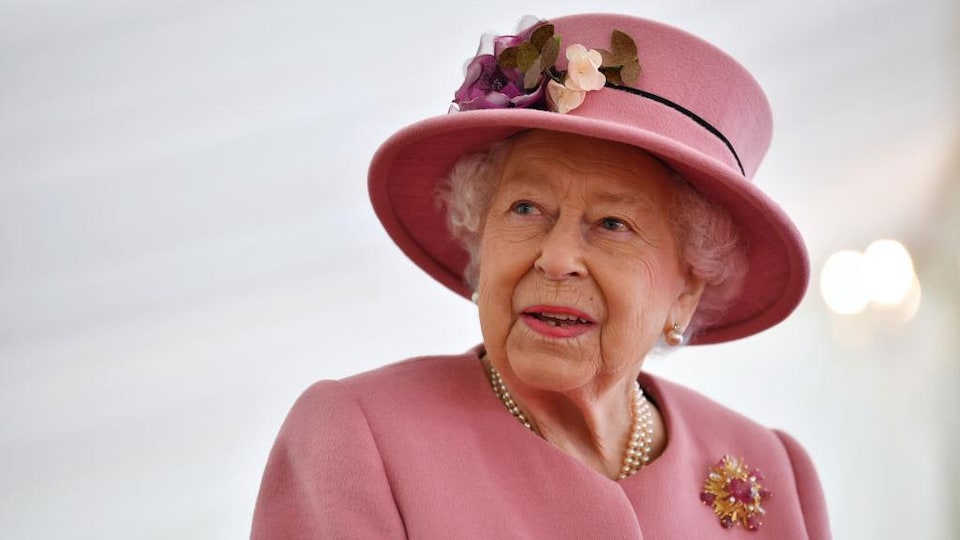 La reine regarde à sa droite. Elle porte un chapeau assorti à sa veste, où figurent une broche de fausses fleurs.