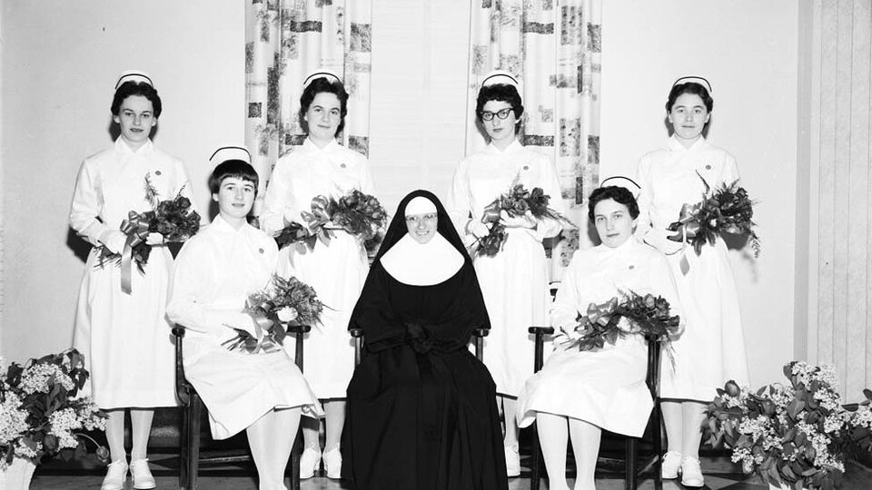 Six jeunes infirmières tenant un bouquet de fleurs entourent une religieuse.