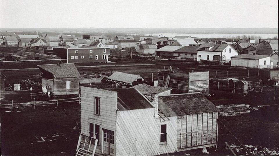 Village sur terre battue avec des bâtisses en construction. Reproduction d'une carte postale.