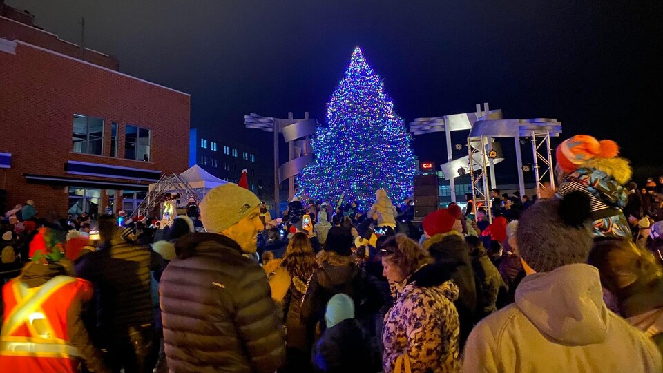 Un sapin de Noël est illuminé sur une place, entouré de personnes. 