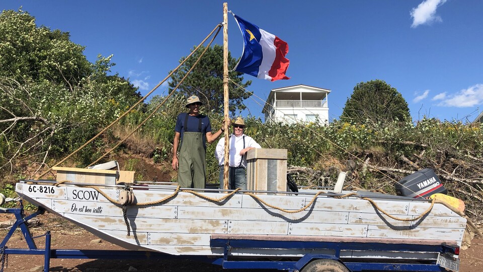 Deux hommes se tiennent debout dans un bateau installé sur une remorque sur le bord d’un rivage.