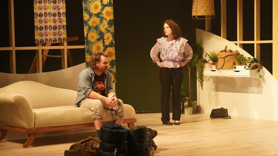 Un homme assis sur un fauteuil et une femme debout dans le décor d'une pièce de théâtre.