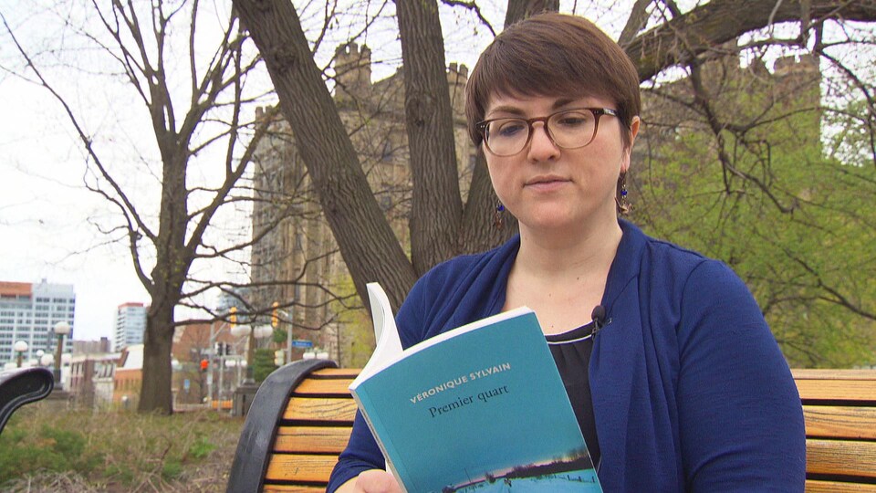 Véronique Sylvain assise sur un banc de parc en train de lire son recueil de poésie « Premier quart ».