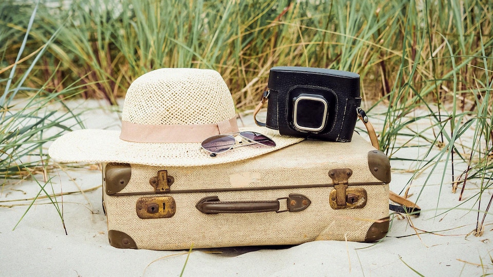 Un chapeau de paille, des lunettes de soleil et un appareil photo sur une valise dans le sable entourée d'herbes hautes.
