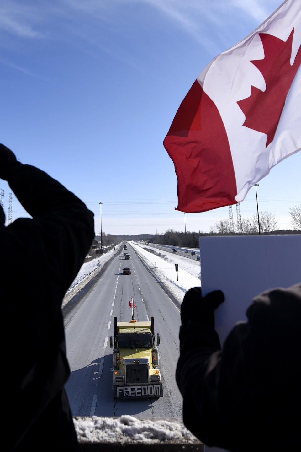 Des manifestants tiennent des drapeaux du Canada du haut d'un viaduc alors qu'un camion passe sur la route en dessous.