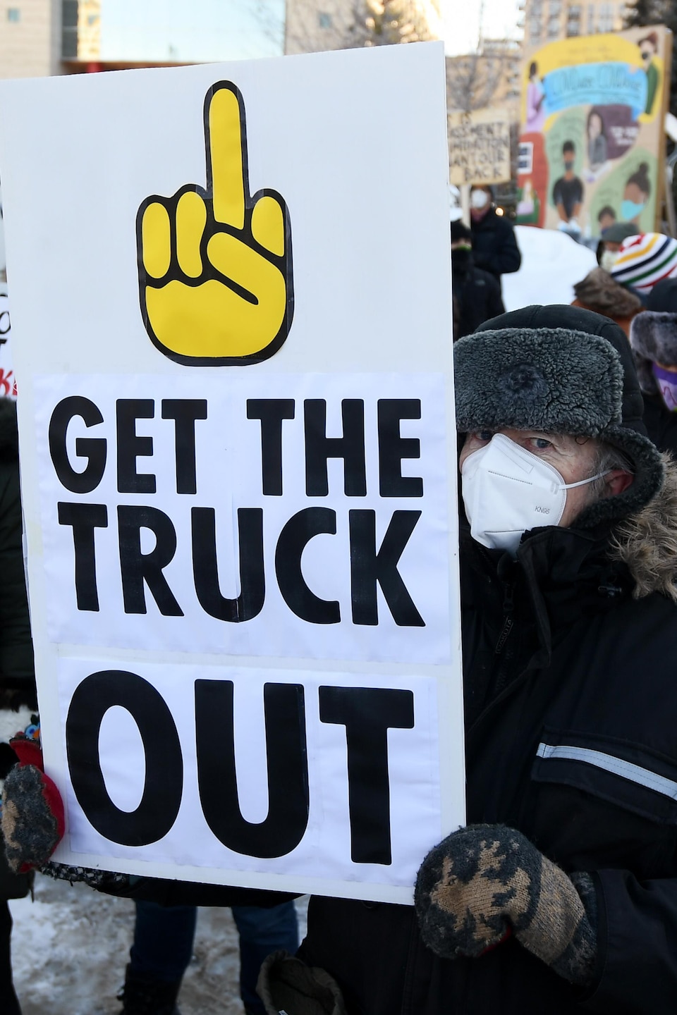 Des personnes masquées tiennent des pancartes devant un immeuble. Sur une des pancarte il est écrit «Get the truck out».