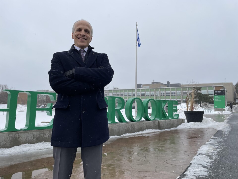 Pierre Cossette se tient avec fierté devant le logo de l'Université de Sherbrooke. 