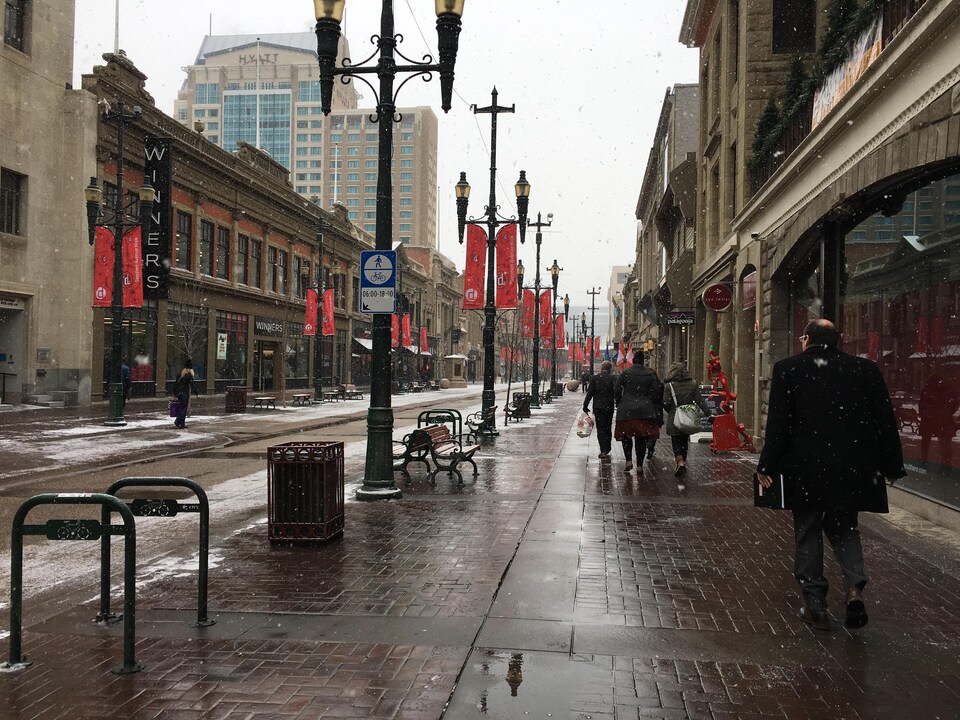 Une rue piétonne au centre-ville de Calgary. De la fine neige tombe. Quelques piétons marchent sur le trottoir, mais la rue est en majorité désertée. 