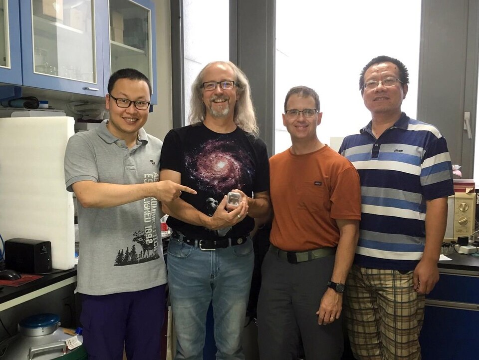 Quatre hommes se tiennent côte-à-côte dans un laboratoire scientifique. L'homme le plus à gauche montre du doigt une petite boîte que tient son acolyte. 