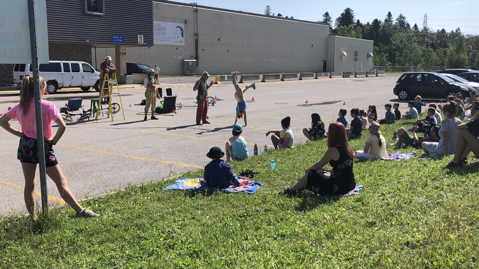 Sous la supervision de moniteurs, des dizaines de jeunes regardent des clowns performer dans un stationnement attenant à un bâtiment public.