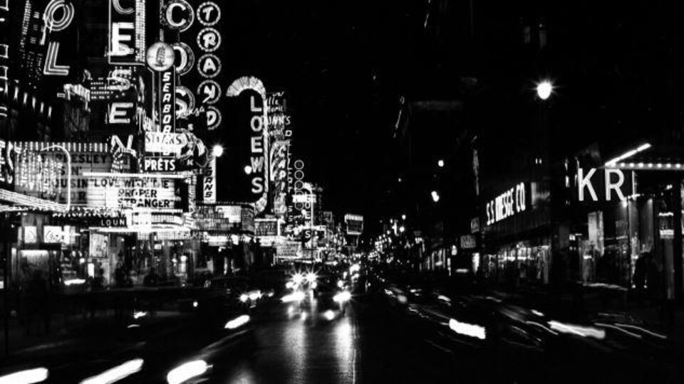 La nuit sur la rue Ste-Catherine avec des voitures et de nombreux éclairages et devantures de cinéma, salles de spectacles et magasins.