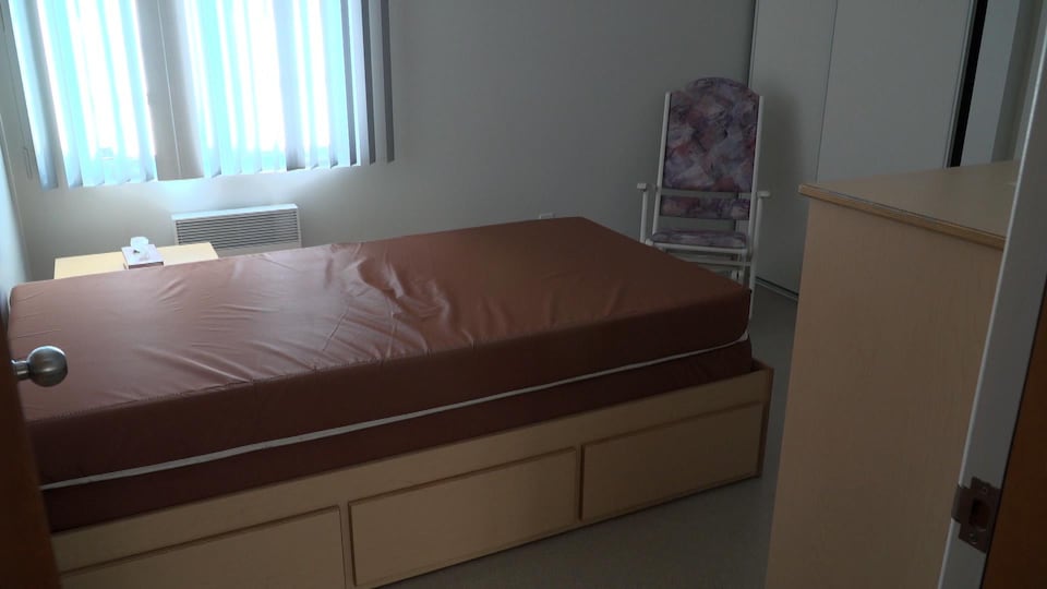Un lit simple sans drap, dans une petite chambre avec une chaise et une commode.