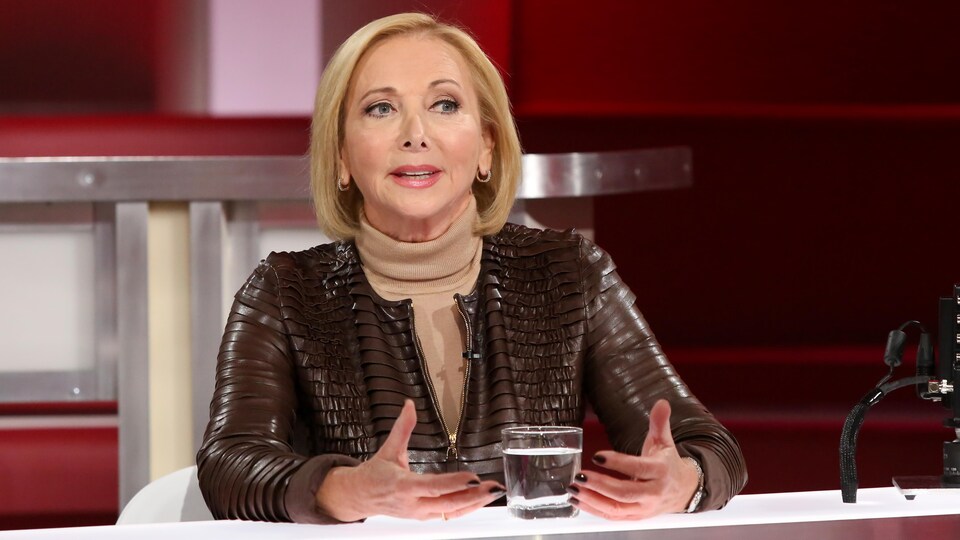 Christiane Germain est à la table des invités. Elle porte un chandail à col roulé beige et une veste en cuir brun.