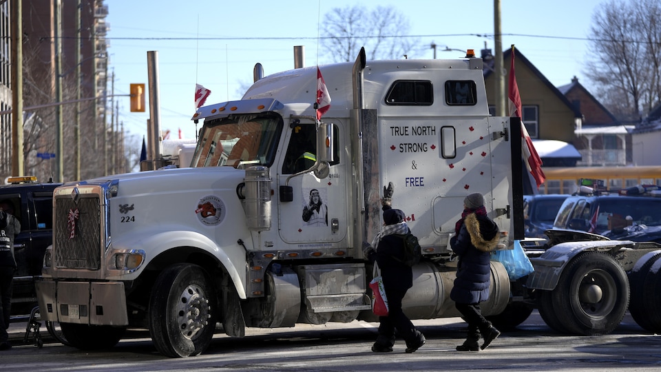 Deux personnes sur une rue saluent une troisième personne dans un camion à 18 roues. Sur le camion, il est écrit: «True North Strong & Free ».