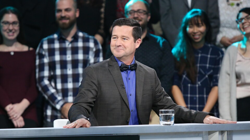 Il porte une chemise bleue, un noeud papillon noir et un veston gris.