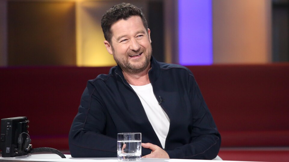 Dany Turcotte sourit. Il porte un t-shirt blanc et un blouson marine. Un verre d'eau est posé sur la table devant lui.
