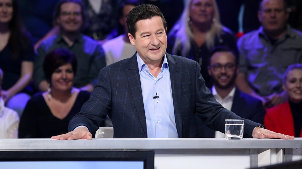 Il sourit. Il porte une chemise bleu pâle et un veston à carreaux.