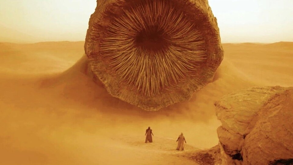 Dans un désert, la bouche d'un immense monstre des sables devant deux silhouettes.