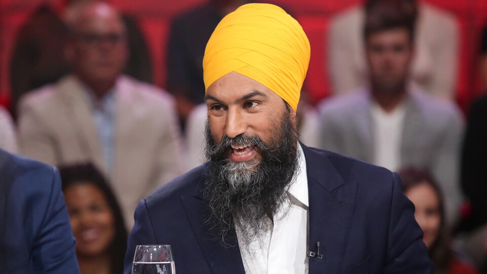 Il porte une chemise blanche, un veston bleu marin et un turban jaune-or. Il porte une barbe.