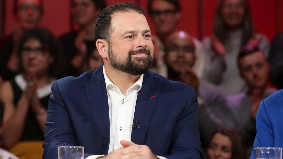 Une homme qui porte une chemise blanche et un veston bleu. Sa barbe et ses cheveux sont bruns.