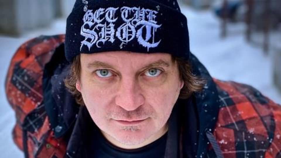 Un homme avec un chapeau fixe l'objectif de la caméra, à l'extérieur l'hiver.