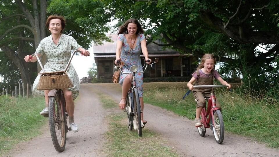Une femme plus âgée, une jeune femme et une petite fille, sur une route de campagne, toutes les trois sur des vélos.
