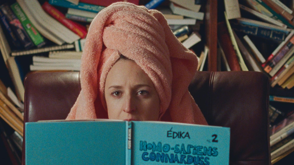 Une femme (Anne-Élisabeth Bossé), une serviette rose sur la tête, lit une bande-dessinée d'Edika.