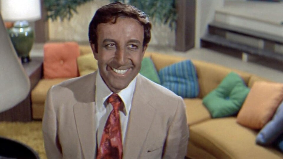 Un homme en costume, souriant, dans un salon.