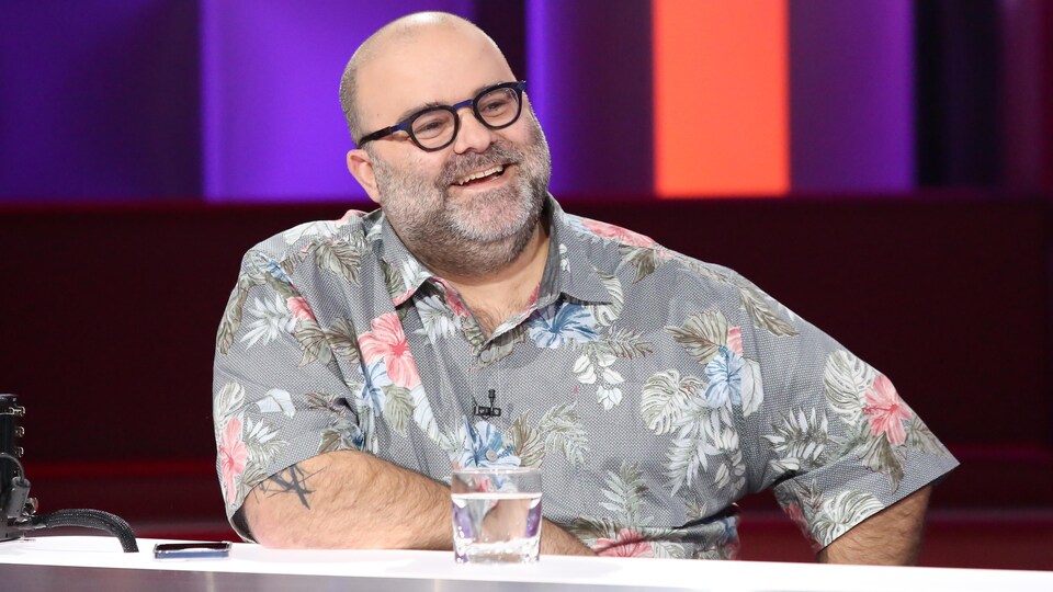 L'homme est assis derrière une table sur un plateau de télévision. Il porte une chemise à fleurs.