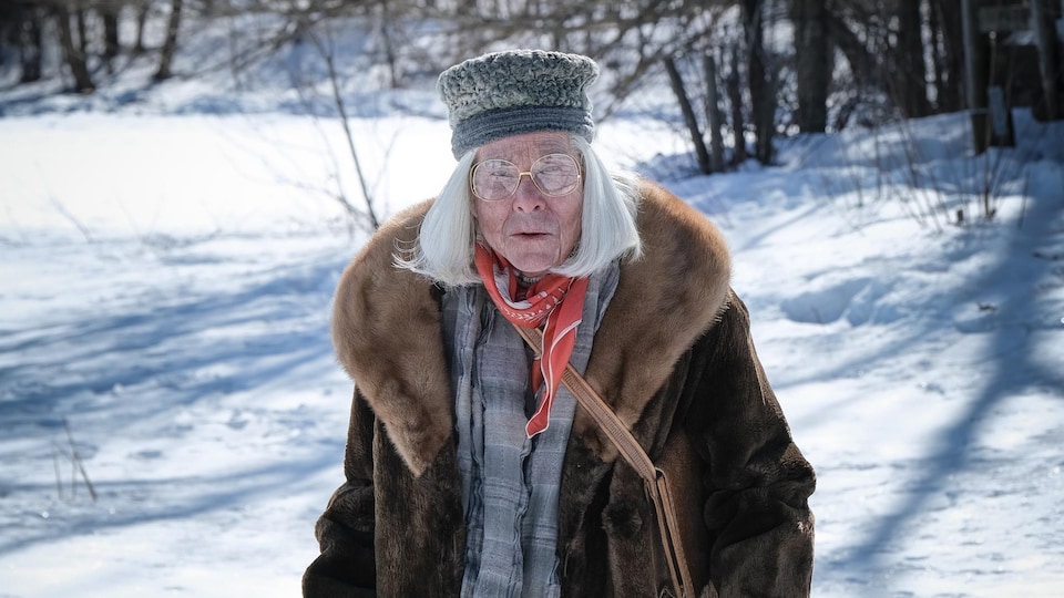 Une vieille femme (Martin Villeneuve) portant des sacs d'épicerie marche dans la neige.