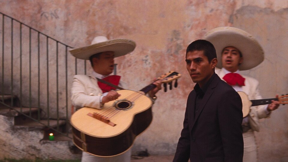 A young man in a black suit walks in front of two mariachi dressed in white.