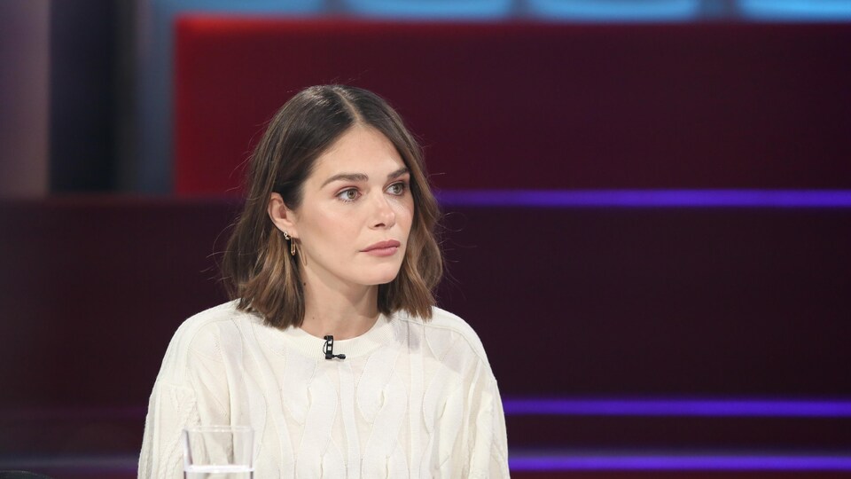 La femme habillée de blanc est assise dans un studio de télévision et regarde l'animateur qu'on ne voit pas.
