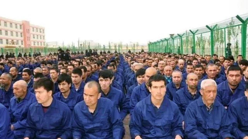 On voit plusieurs prisonniers ouïgours assis en rang dans un camp d'internement.