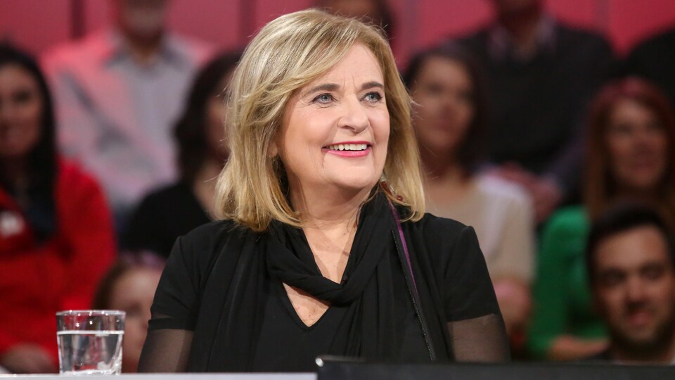 Une femme aux cheveux blonds mi-longs. Elle sourit. Elle porte une robe noire.