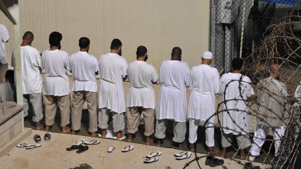 Des prisonniers, en blanc, de dos, en train de prier.