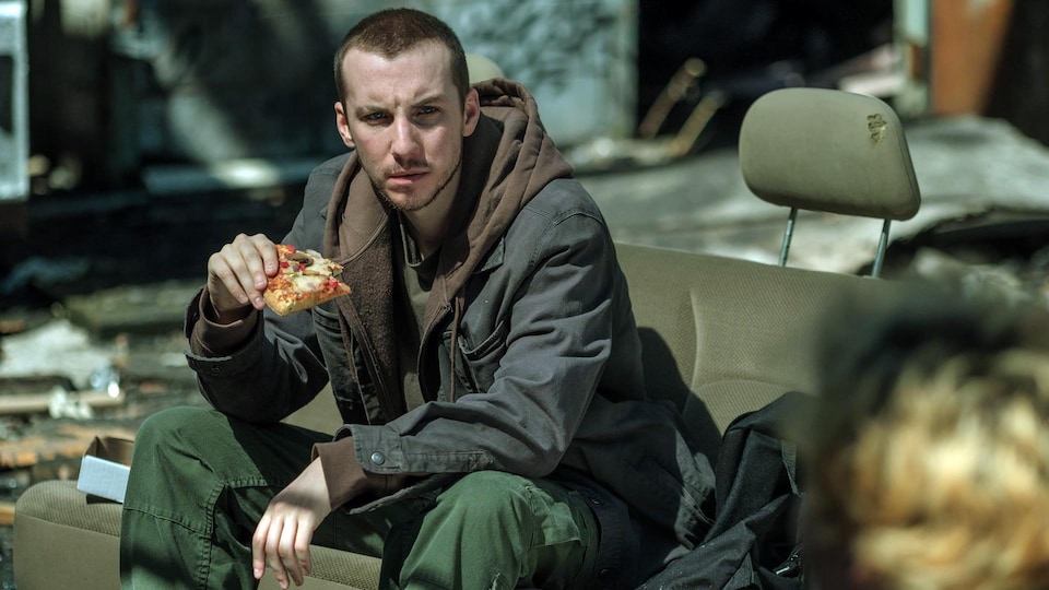 Jessy est assis sur un vieux siège de fourgonnette, dans un lieu abandonné et il mange une pizza.