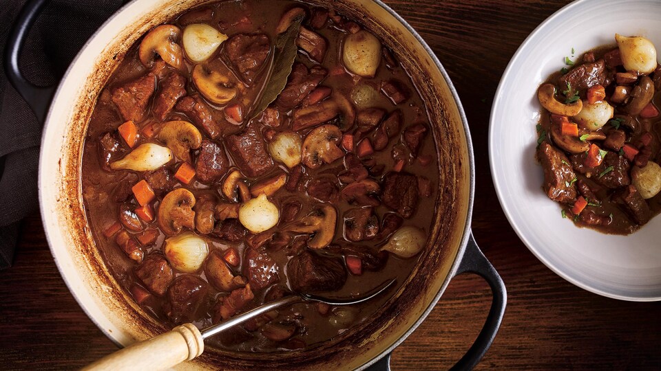 Un chaudron rempli de boeuf bourguignon sur une table, à côté d'une assiette qui en contient une portion.