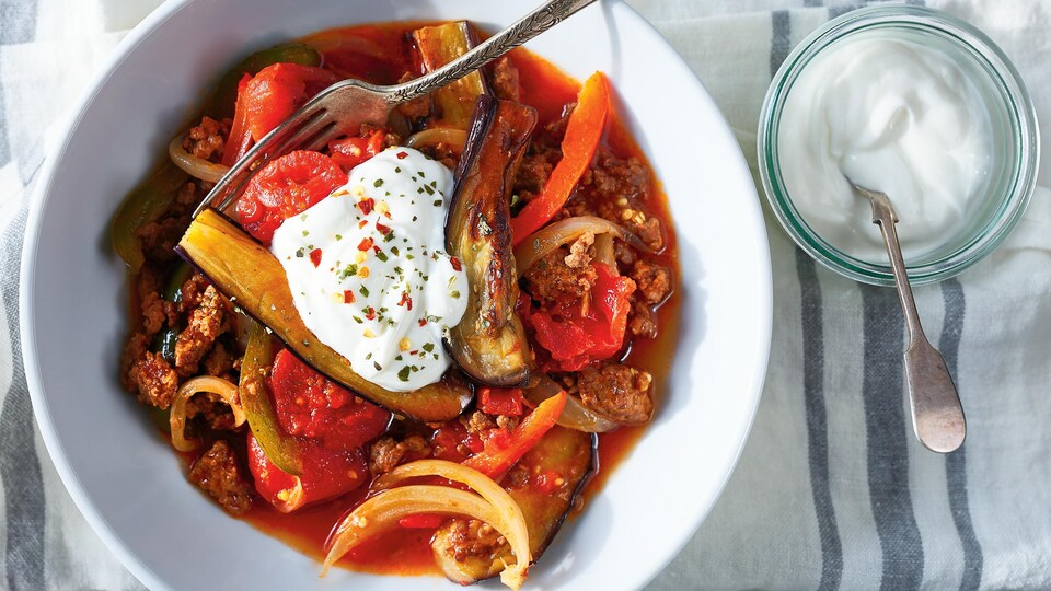 Un bol de ragoût d'aubergine à la viande qui contient des tomates, des oignons et des poivrons. Le plat est accompagné de yogourt nature, parsemé de flocons de piments et d'origan.
