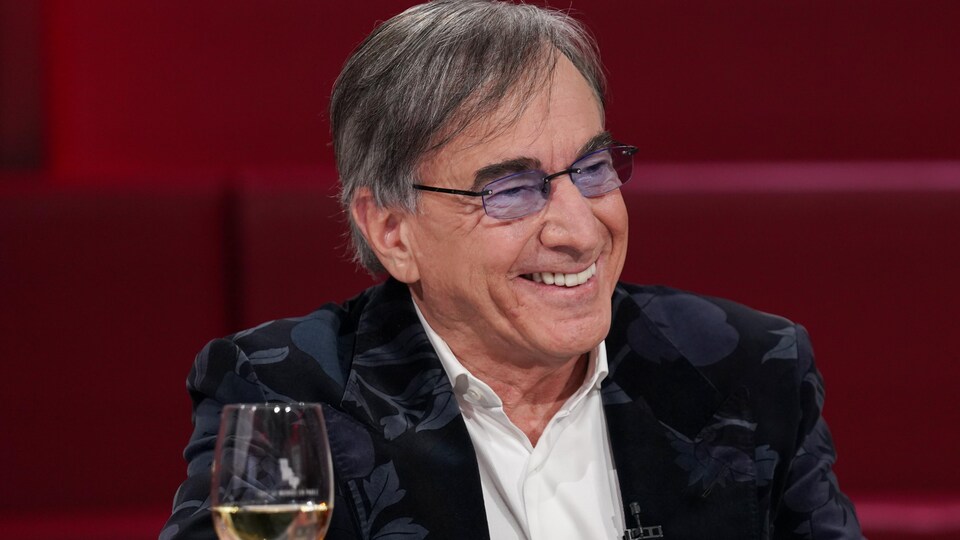 L'homme est assis dans un studio de télévision. Il sourit.