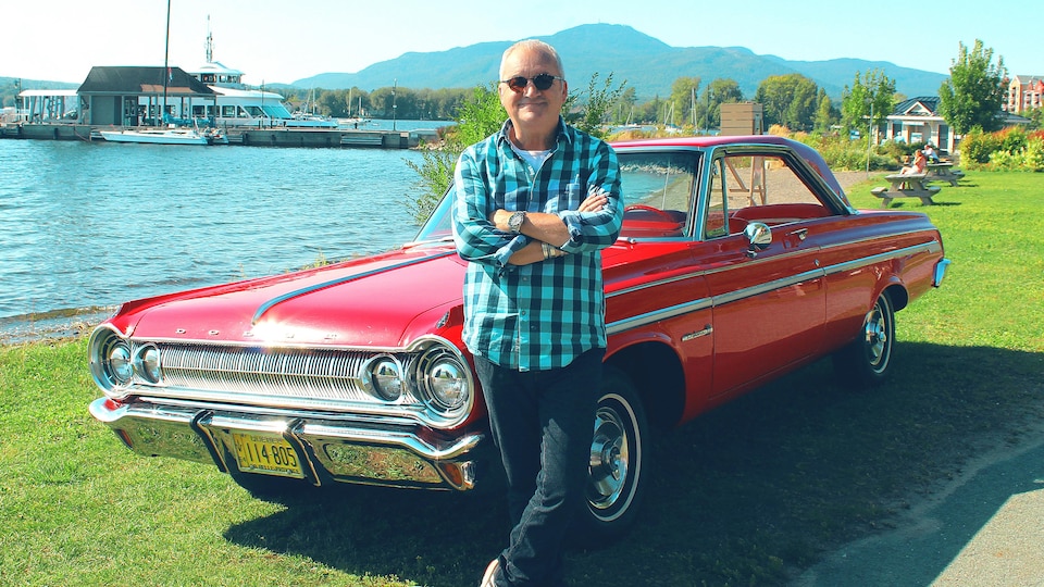 Michel Barrette est assis, accoté sur le capot d'une vieille voiture rouge.