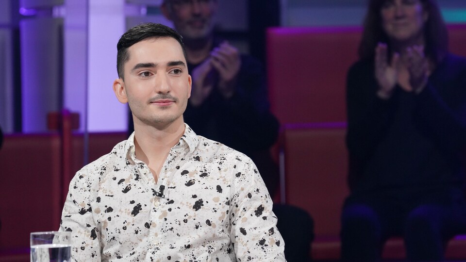 Il est sur le plateau de Tout le monde en parle, porte une chemise blanche tachetée, sourit très légèrement.