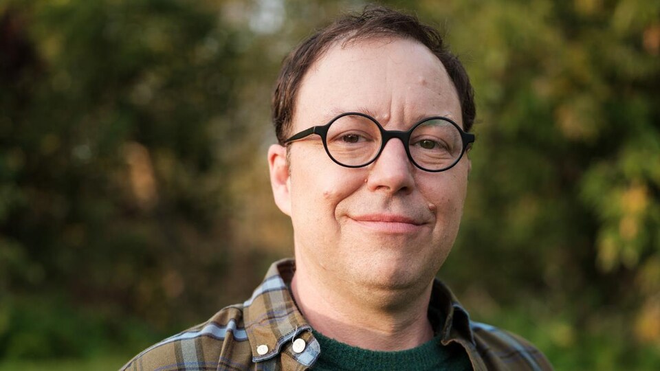 Le comédien est photographié dehors et sourit en regardant l'objectif. Il porte des lunettes et une chemise à carreaux. 