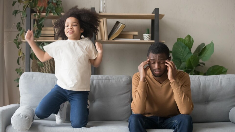 Père afro-américain ayant des problèmes avec une fille préscolaire coquine et bruyante. Elle saute sur un canapé et exige de l'attention. Le père fatigué se touche les temples.