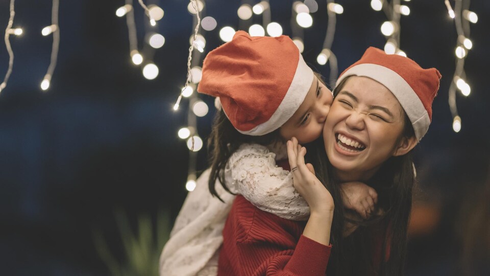 Une maman et sa fille, d'origine asiatique, portent des bonnets de Noel et rient en se faisant un câlin